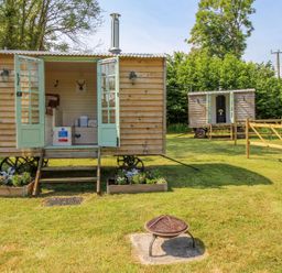 Two wooden shepherd huts on wheels with open double doors on a grassy area with flower beds and a fire pit at Acres Meadow in Maiden Newton