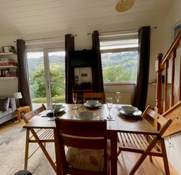 A dining area with a wooden table set for four next to a window and glass door with curtains in a room with a staircase and a sofa