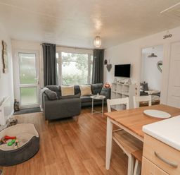 A living room with a gray sofa and pillows a small white table a wall-mounted TV open doorway to a bedroom dining table and chairs a dog bed with toys on a wooden floor at Number 24 The Park in Kilkhampton