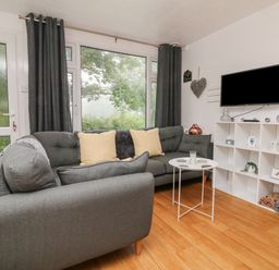 A living room with a gray sofa and yellow cushions a white storage unit with a television and a small white round table at Number 24 The Park Kilkhampton