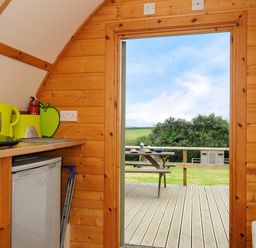 A wooden kitchen interior with a small refrigerator kettle toaster and fire extinguisher with a door opening to a deck with a picnic table and green landscape at Sheep Shed Penbugle Organic Farm St Keyne near Looe