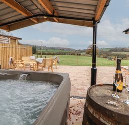 An outdoor hot tub under a wooden shelter with wooden chairs and table on a patio overlooking a green field at Elliw in Bontuchel near Ruthin