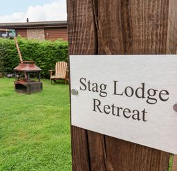 A grassy outdoor area with a fire pit, wooden chair, swing set, and a sign on a wooden post reading Stag Lodge Retreat