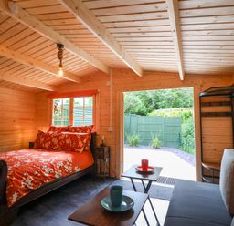 A wooden interior room with a bed with red floral bedding a small side table a gray sofa two small tables with cups and an open doorway showing a fenced garden at Log Cabin - Amber Glow in Glastonbury