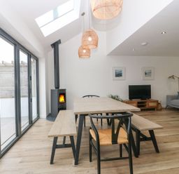 A dining area with a wooden table and benches next to a living room with a gray sofa and a TV at 11 Capel Farm in Trearddur Bay
