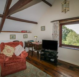 A room with a red armchair a wooden desk with a lamp a chair a television on a glass stand and a window showing trees at Beckaford Cottage in Bovey Tracey
