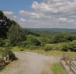 An outdoor scene with a gravel path flowers trees and a wooden fence with hills and clouds in the background at Beckaford Cottage in Bovey Tracey