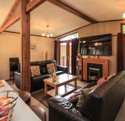 A living room with black leather sofas a wooden coffee table a TV mounted above a fireplace and a fruit basket on a counter at Templar Rise 16 - Devon Comfort Finlakes Holiday Park near Chudleigh