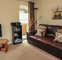 A living room with a brown leather sofa with cushions a wooden coffee table a bookshelf with books and a stereo a tall floor lamp and a wall picture of a boat at The Brambles 17 - Conkers Lodge at Finlakes Holiday Park near Chudleigh
