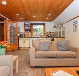 A living room with a sofa and two cushions a wooden coffee table with a remote and paper a kitchen area with cabinets and appliances a wooden ceiling and a vase with flowers Lodge 45 - Lazy Bear Finlakes Holiday Park near Chudleigh
