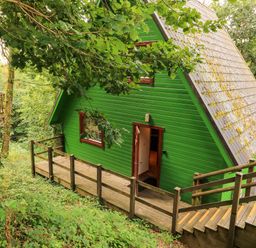 A green wooden A-frame cabin with a wooden deck and stairs surrounded by trees at Lodge 58 - Oak Tree Lodge in Finlakes Holiday Park near Chudleigh