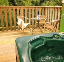 An outdoor deck with a hot tub a table with two chairs and a bottle with glasses at Lodge 58 - Oak Tree Lodge in Finlakes Holiday Park near Chudleigh