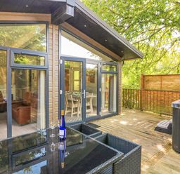 An outdoor deck with a glass table set a wine bottle and two glasses a hot tub and patio doors leading to the living area at Woodland View 7 - Oak Cabin Finlakes Holiday Park near Chudleigh