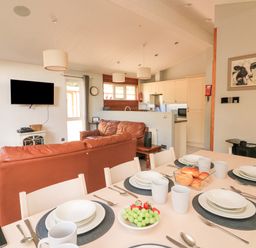 A dining table set with plates cups and cutlery in an open plan kitchen and living area with brown leather sofas and a wall mounted TV at Woodland View 7 - Oak Cabin Finlakes Holiday Park near Chudleigh