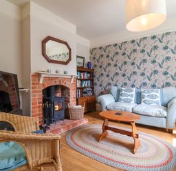 A living room with a blue sofa, wicker chair, wooden coffee table, brick fireplace, and patterned wallpaper at Samphire House in Southwold