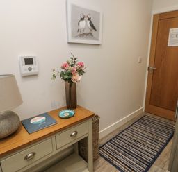 A hallway with a striped rug a wooden door a table with drawers a lamp a vase with flowers and a guest book Sandy Bay Retreat Benllech