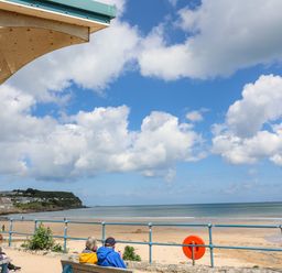 A beach view with people sitting on benches near a railing under a partly covered structure at Sandy Bay Retreat in Benllech