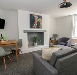 A living room with a gray sofa, two chairs, a small wooden dining table with two chairs, a wall-mounted television, a fireplace with a plant inside, and a colorful giraffe painting above at Flat 1 - Sandbanks in Bournemouth