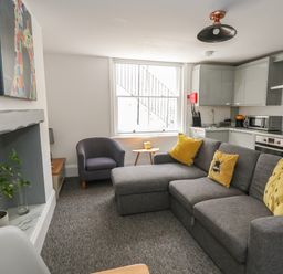 A living room with a grey sofa, yellow cushions, a grey armchair, a small wooden table, a fireplace with a plant, and a kitchen area at Flat 1 - Sandbanks in Bournemouth