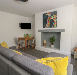 A living room with a gray sofa with yellow cushions a wooden table with two gray chairs a television mounted on the wall a decorative fireplace with a plant a candle and a vase and a painting of colorful giraffes above the fireplace at Flat 1 - Sandbanks in Bournemouth
