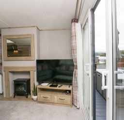A living room with a television on a wooden stand, an electric stove in a fireplace, a mirror above, curtains on windows, and a sliding glass door leading to an outdoor seating area at Mon in Troutbeck Bridge