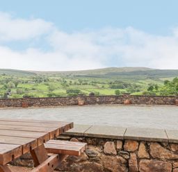 A wooden picnic table on a stone patio overlooking a stone wall and green hills at Llys-y-Wennol in Penmachno