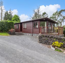 A gravel driveway leading to a wooden lodge with a stone base and a small deck with outdoor furniture surrounded by trees and shrubs at Lodge 3 At Pound Farm in Crook near Kendal