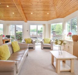 A living room with beige sofas and yellow pillows a wooden coffee table with flowers a wooden ceiling and large windows at Lodge 3 At Pound Farm in Crook near Kendal