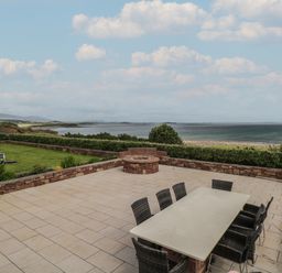 An outdoor patio with a dining table and chairs next to a fire pit and a grassy area with a barbecue overlooking the sea at Cois Leice Cushlecka near Mulranny County Mayo