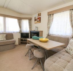 A living area with sofas a wooden table two stools a television and windows with curtains at 23 Manleigh Park in Combe Martin