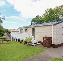 A beige mobile home with white railings and stairs on a lawn with a wooden picnic table and trees at 23 Manleigh Park in Combe Martin