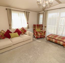 A living room with a beige sofa with cushions a plaid armchair and ottoman with curtains and windows at The Boathouse in East Wittering