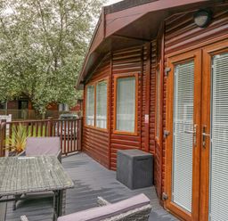 An outdoor wooden deck with a table, two chairs, a storage box, and a wooden lodge with windows and glass doors at Don's Lodge in Grasmere 3 White Cross Bay near Windermere