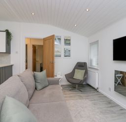 A living room with a gray sofa, armchair, wall-mounted television, and open wooden door at Don's Lodge in Grasmere 3 White Cross Bay near Windermere