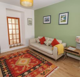 A living room with a beige sofa and colorful cushions a wooden side table with a lamp a patterned rug and bookshelves at Bryn Siriol in Narberth