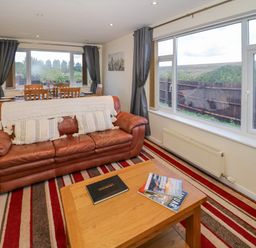A living room with a leather sofa and wooden coffee table next to large windows at Fenny Marina Cottage in Fenny Bentley