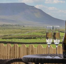 A view of a mountain and green fields with sheep behind a wooden fence with a table holding two champagne glasses and a bottle at Lapwing in Chapel Le Dale near Ingleton