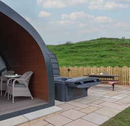 An outdoor patio area with two wicker chairs and a small table under a curved wooden structure a picnic table with items on it a closed hot tub a chiminea and a wooden fence with grassy hills in the background at Lapwing in Chapel Le Dale near Ingleton