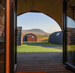 View from inside a wooden pod with open glass doors showing patio chairs lawn path and other pods with a mountain in the background at Oyster Catcher in Chapel Le Dale near Ingleton
