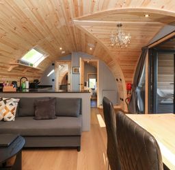 A curved wooden interior with a gray sofa and cushions a black coffee table a wooden dining table with black chairs a chandelier and glass doors leading to an outdoor seating area at Oyster Catcher in Chapel Le Dale near Ingleton