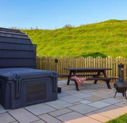 An outdoor patio with a covered hot tub a wooden picnic table with a blanket and a metal chiminea at Heron in Ingleton