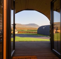 View through open glass doors onto a wooden porch lawn a curved gray structure and a mountain in the distance at Heron in Ingleton