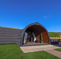 A small modern curved cabin with a patio, chair, and plant on grass at Wren Ingleton