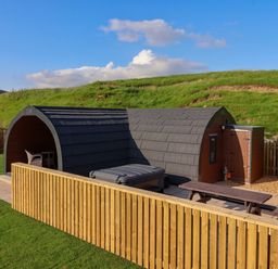 An arched wooden cabin with tiled roof surrounded by a wooden fence and grassy hills in the background at Wren in Ingleton