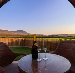 A round glass table with a bottle and two glasses between two wicker chairs on a covered patio overlooking a fenced grassy yard and distant hills at Barn Owl in Chapel Le Dale near Ingleton