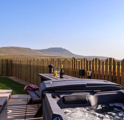 An outdoor area with a hot tub a wooden picnic table with drinks and glasses and a wooden fence with hills in the background at Barn Owl in Chapel Le Dale near Ingleton