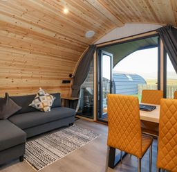 A wooden interior living space with a dark grey sofa and patterned cushions near orange quilted dining chairs around a table at Barn Owl in Chapel Le Dale near Ingleton
