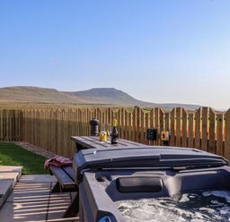 An outdoor area with a hot tub next to a picnic table with drinks and glasses and a wooden fence in the background at Curlew in Chapel Le Dale near Ingleton