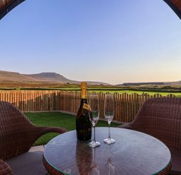 A patio with two wicker chairs around a glass table with a bottle of sparkling wine and two wine glasses overlooking fields and hills at Curlew in Chapel Le Dale near Ingleton