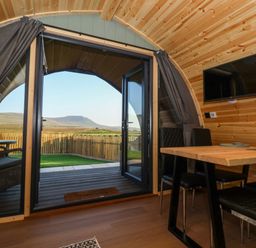 Interior of a wooden pod with a dining table and chairs next to double glass doors opening to a fenced yard and distant hills at Curlew in Chapel Le Dale near Ingleton